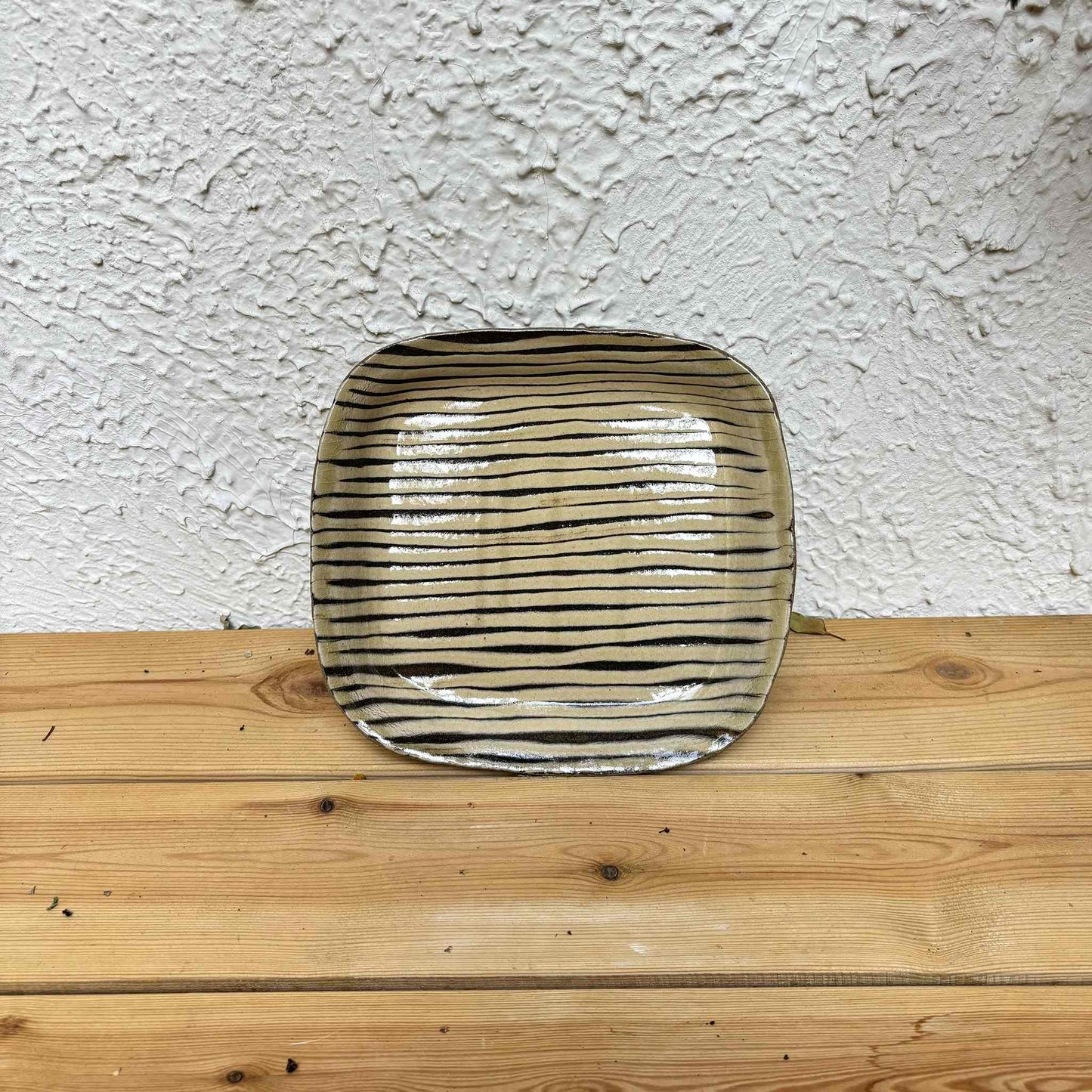 Handmade square plate with unique, hand-applied surface patterns in a neutral, earthy tone, set against a natural wood background.