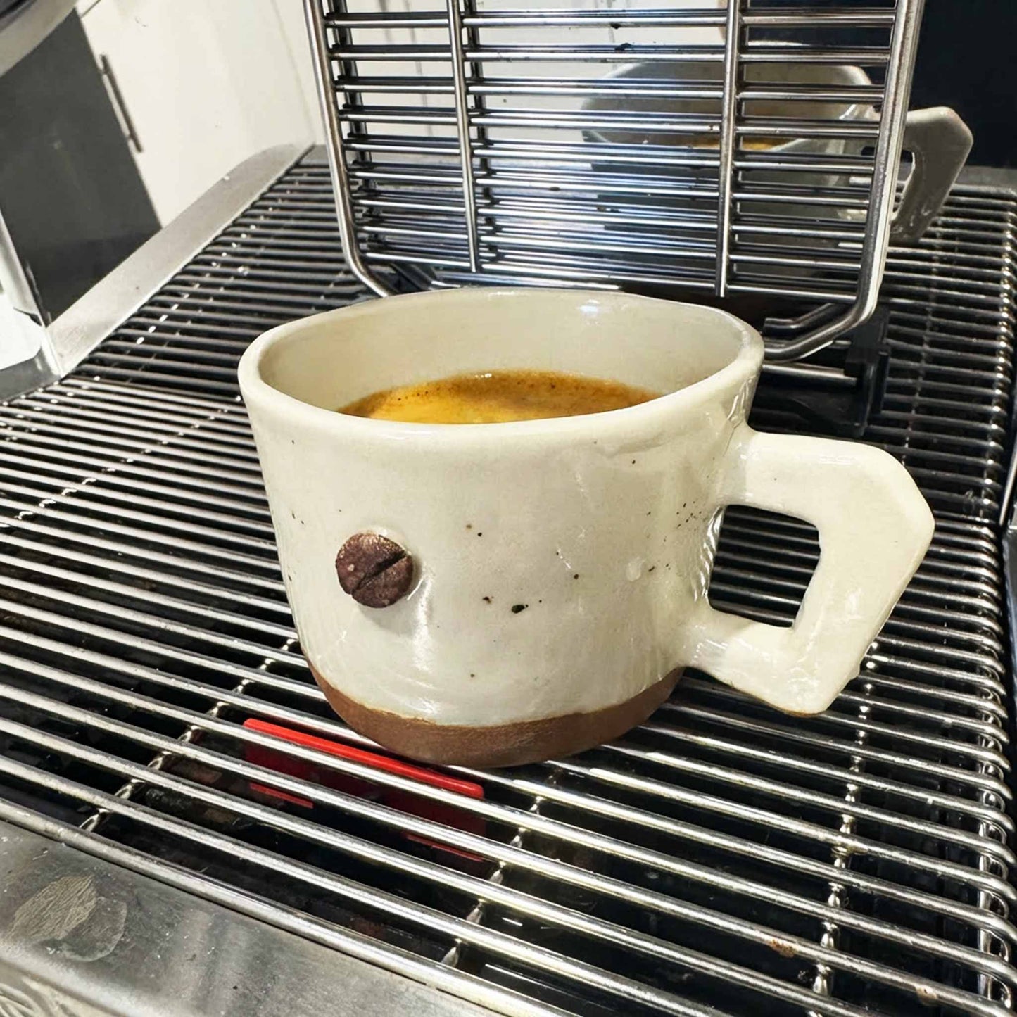 Handmade white ceramic espresso cup with a brown handle, sitting on a metal grate.