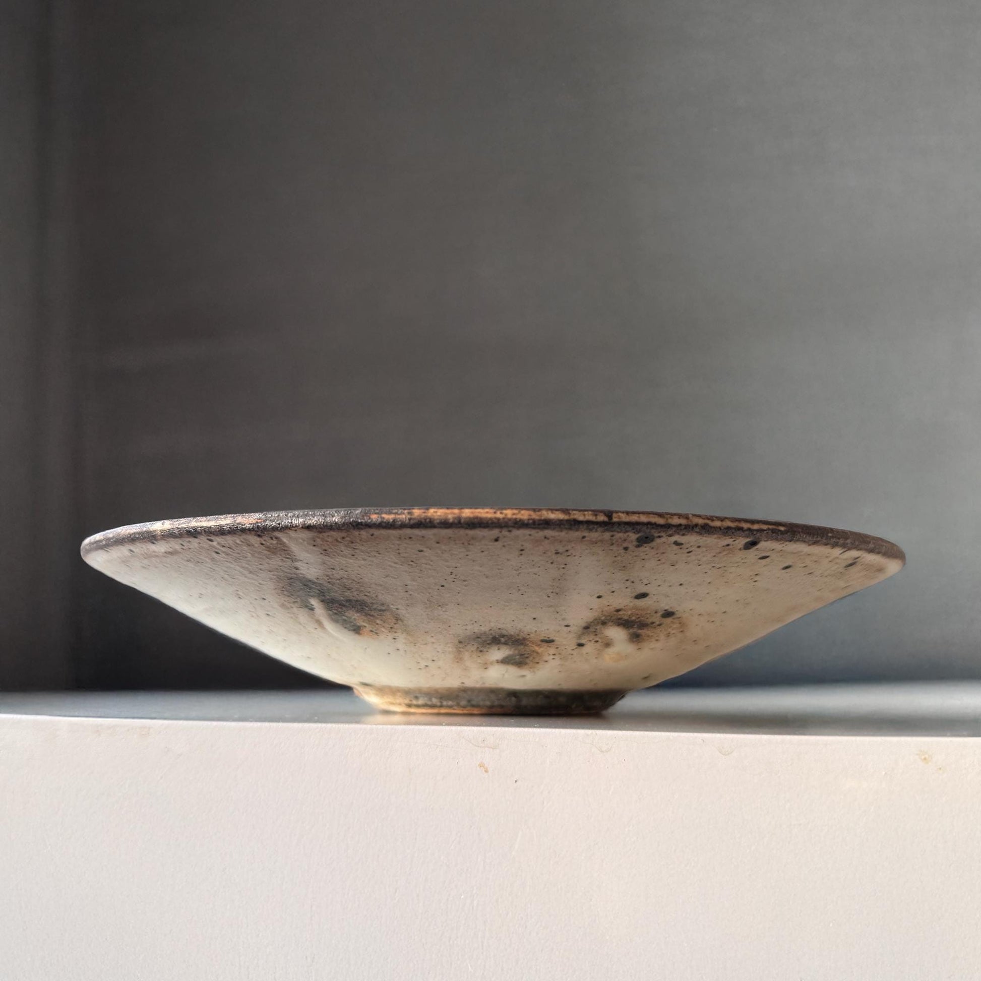 A handcrafted, shallow ceramic bowl with a subtle, earthy tone and a speckled glaze, evoking a sense of understated beauty.