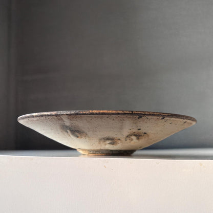 A handcrafted, shallow ceramic bowl with a subtle, earthy tone and a speckled glaze, evoking a sense of understated beauty.