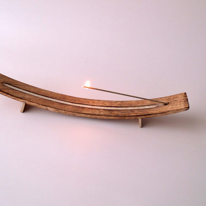 Wooden incense holder with a lit incense stick on a light pink background