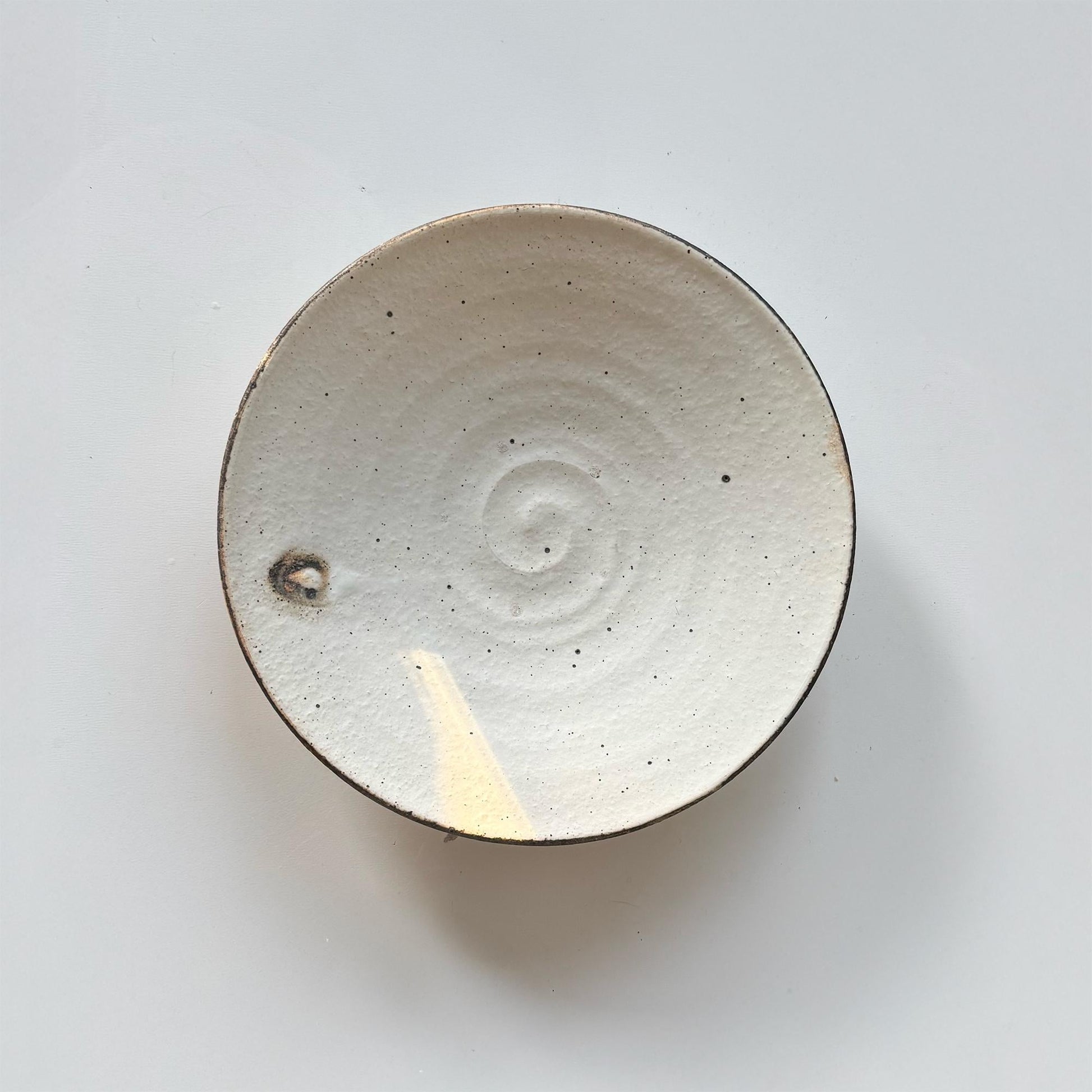 A handcrafted, shallow ceramic bowl with a subtle, off-white glaze and a small, circular indentation in the center.