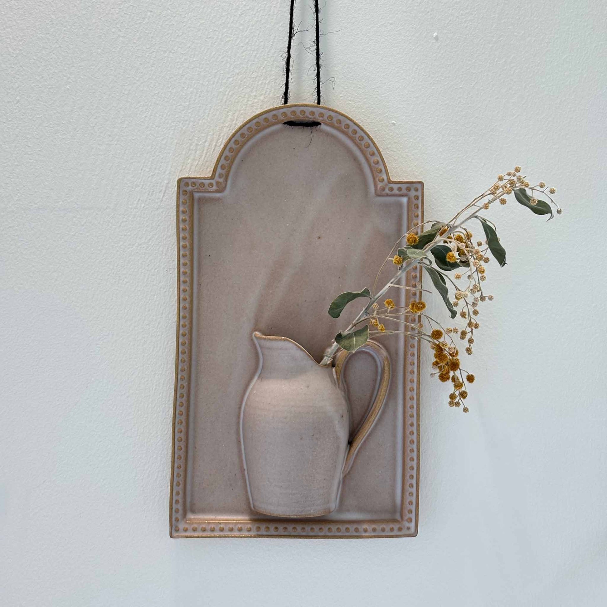 A white, hand-painted wall vase with a deep, earthy tone, suspended from a black string, against a white background.