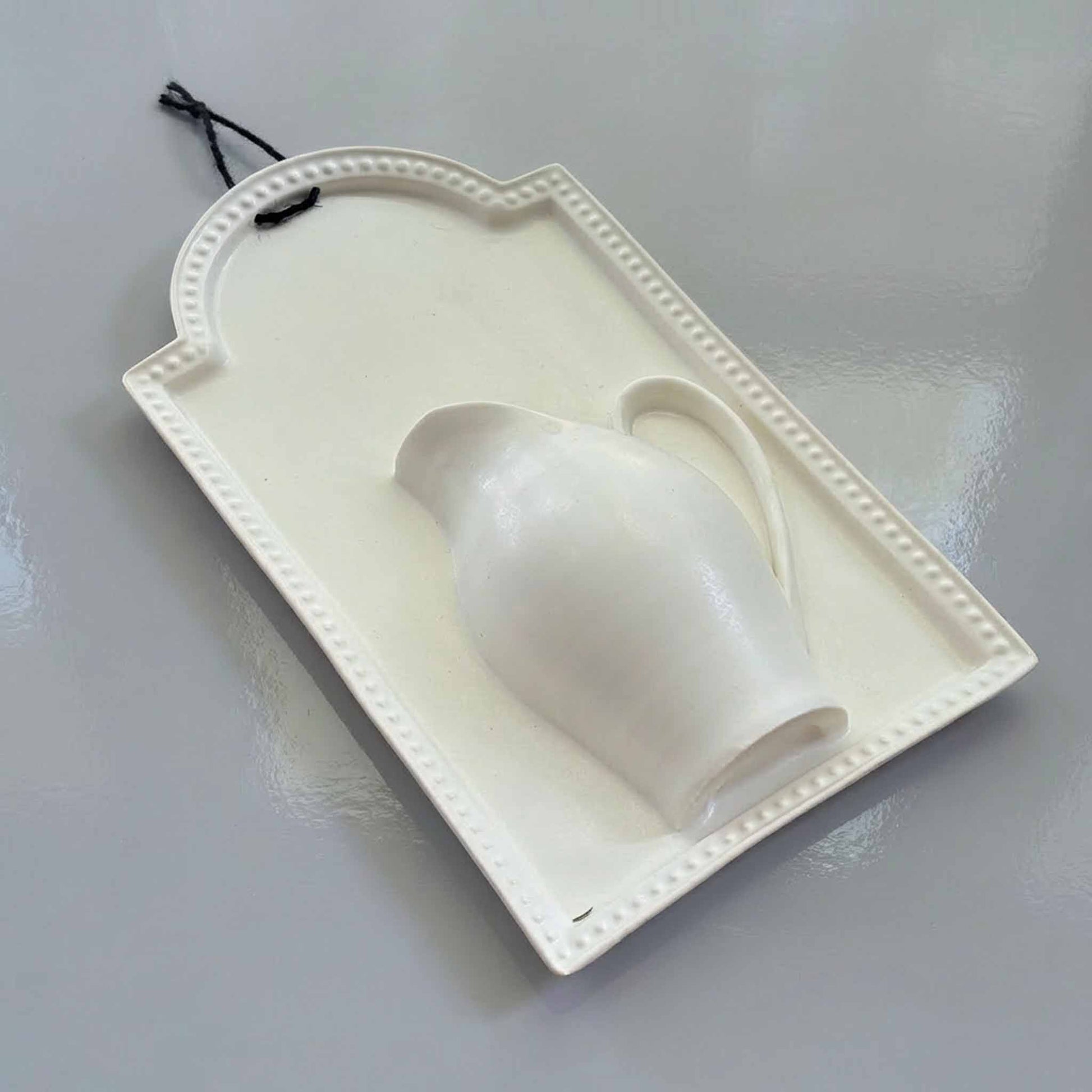 A white, hand-painted wall vase with a delicate handle, suspended from a black string, against a neutral background.