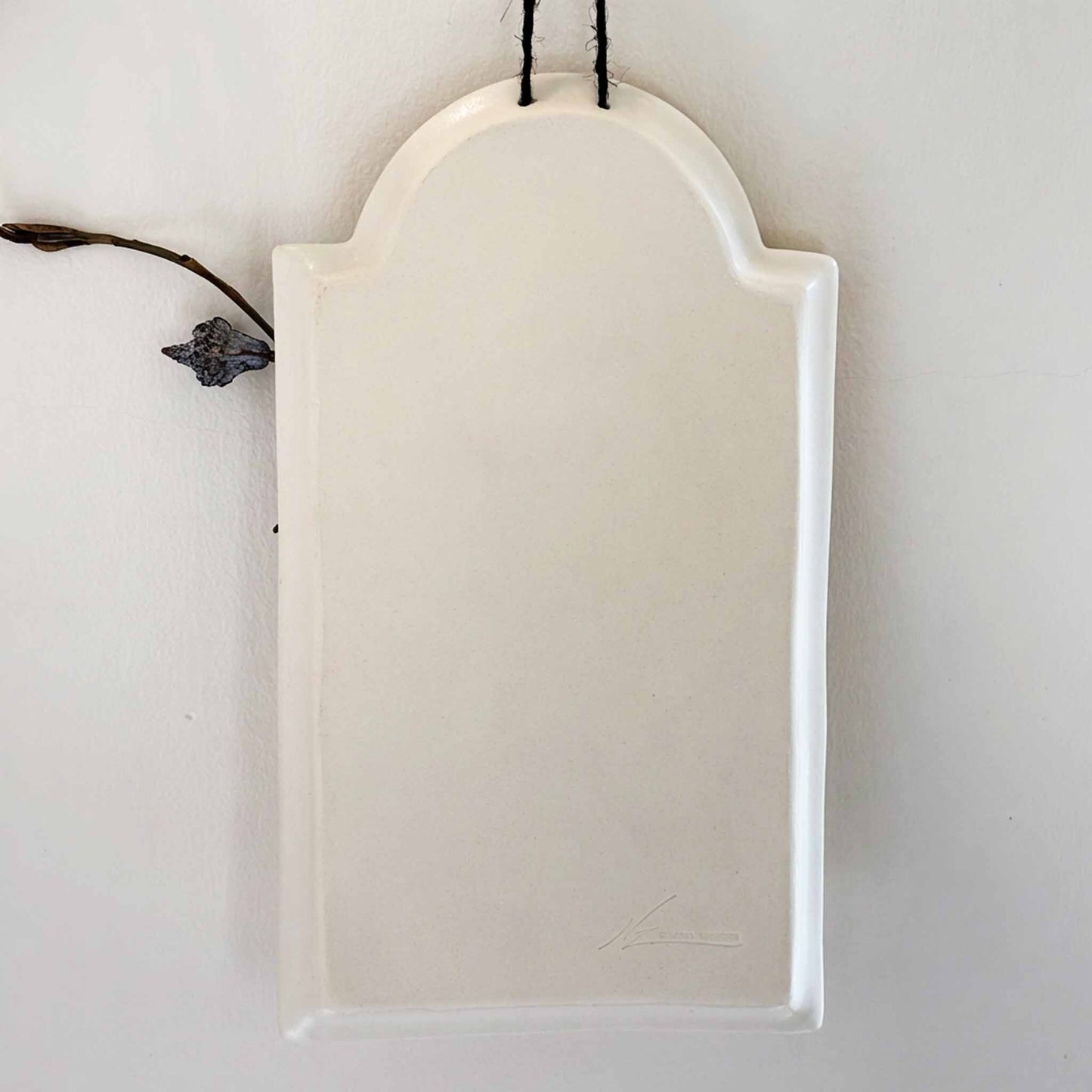 A white, handcrafted wall vase with a delicate, curved design, suspended from a black string against a neutral background.
