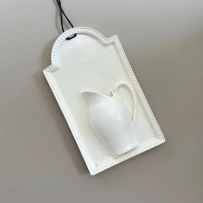 A white, hand-poured wall vase with a delicate, curved design, suspended from a black cord against a neutral background.