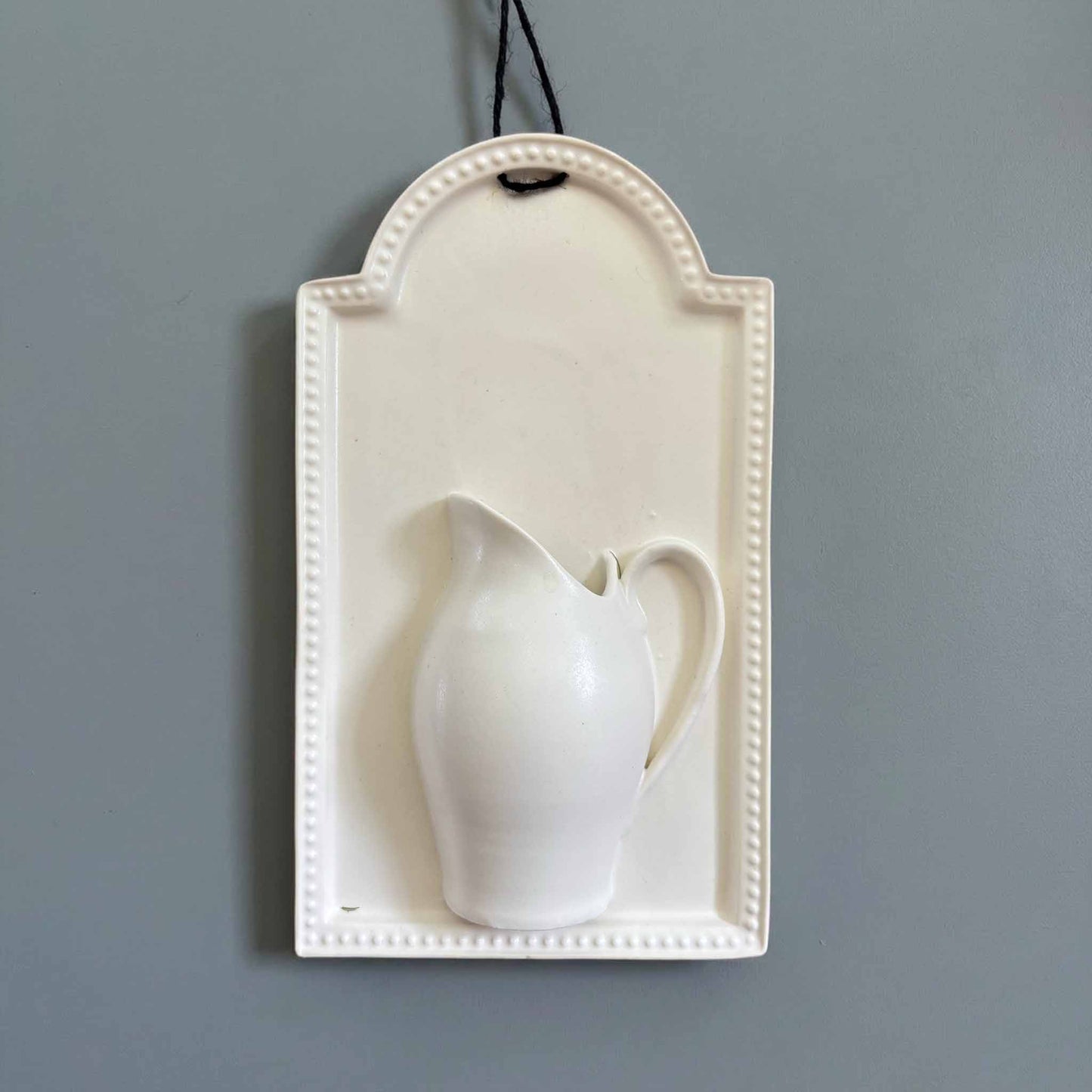 A white, hand-painted wall vase with a delicate, curvaceous design, suspended from a wall by a thin black cord.