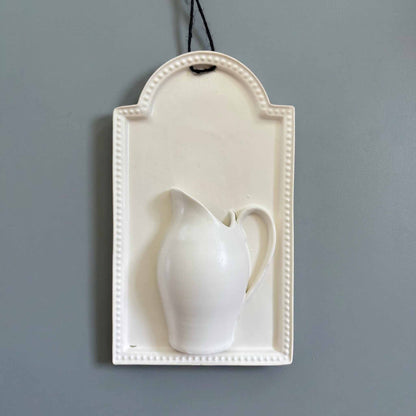 A white, hand-painted wall vase with a delicate, curvaceous design, suspended from a wall by a thin black cord.