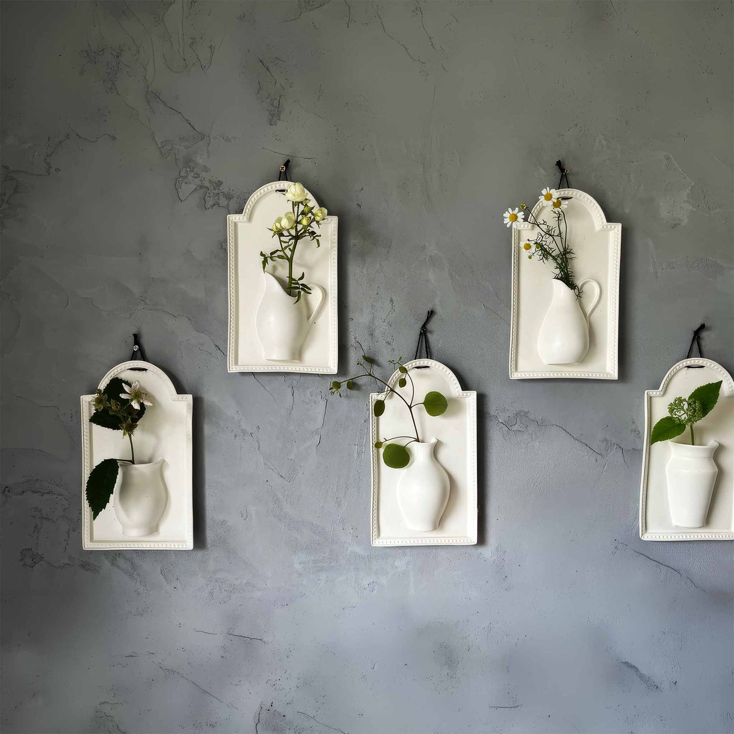 A white, handcrafted wall vase with a delicate, curvaceous design, suspended from a wall by a black metal hook.