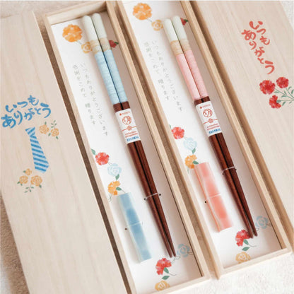 A gift set of chopsticks presented in a light wood box with floral illustrations and Japanese text. The chopsticks have white and pale blue striped handles, dark brown wooden shafts, matching light blue and pink protective sleeves, and decorative paper bands with red and white logos.