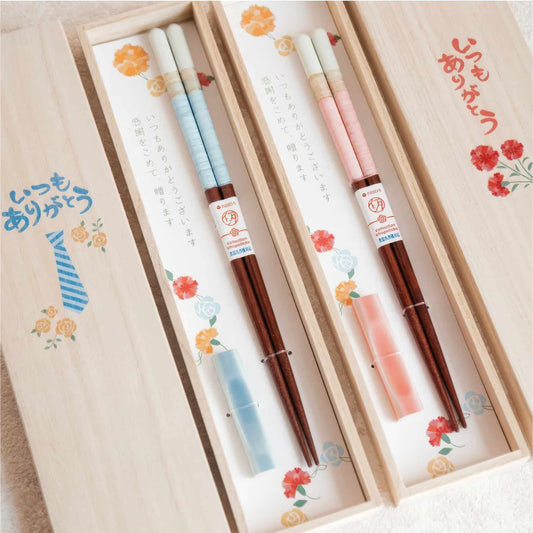 A gift set of chopsticks presented in a light wood box with floral illustrations and Japanese text. The chopsticks have white and pale blue striped handles, dark brown wooden shafts, matching light blue and pink protective sleeves, and decorative paper bands with red and white logos.