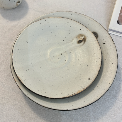 A handcrafted, shallow bowl made of ceramic material, characterized by its light gray color and speckled glaze.